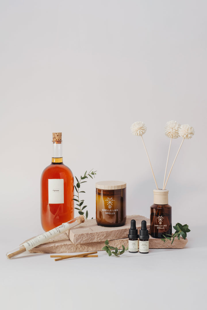 Amber glass home fragrance products, a scented candle, oils and diffuser stems arranged with greenery on a pale background.
